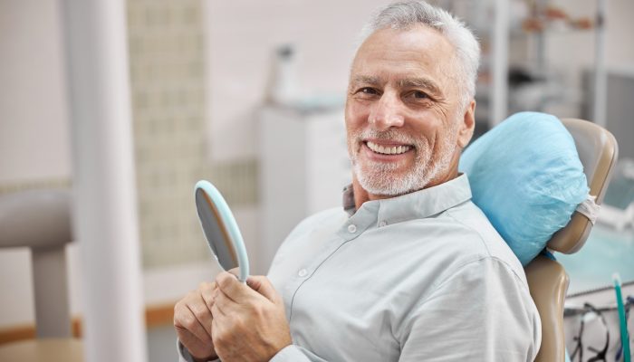 Cheerful patient sitting in a dental chair and holding a mirror while smiling at the camera