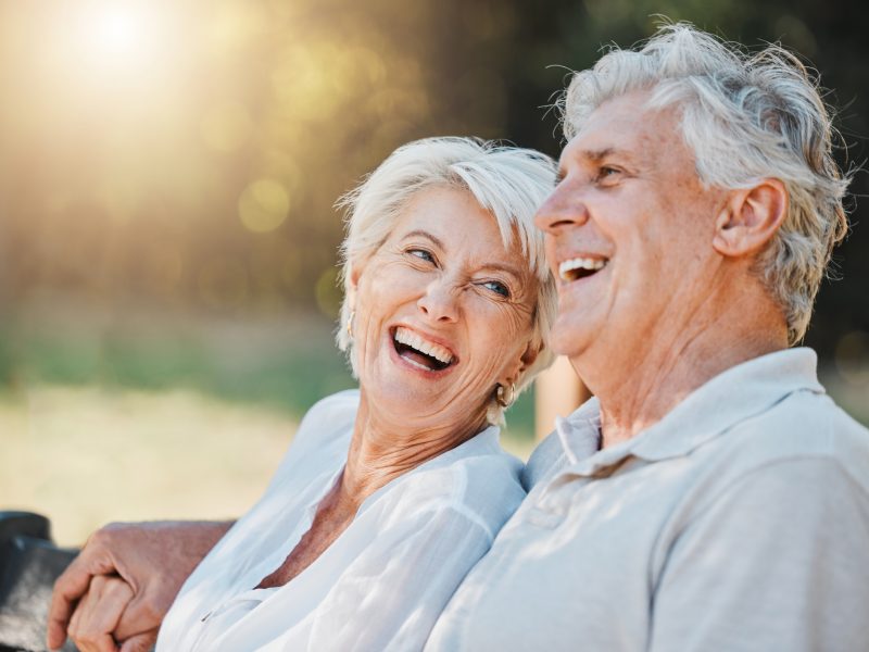Senior couple, laughing or park bench in nature garden for love, support or bonding retirement trust. Smile, woman or elderly man in backyard for joke, funny news or relationship on lens flare space