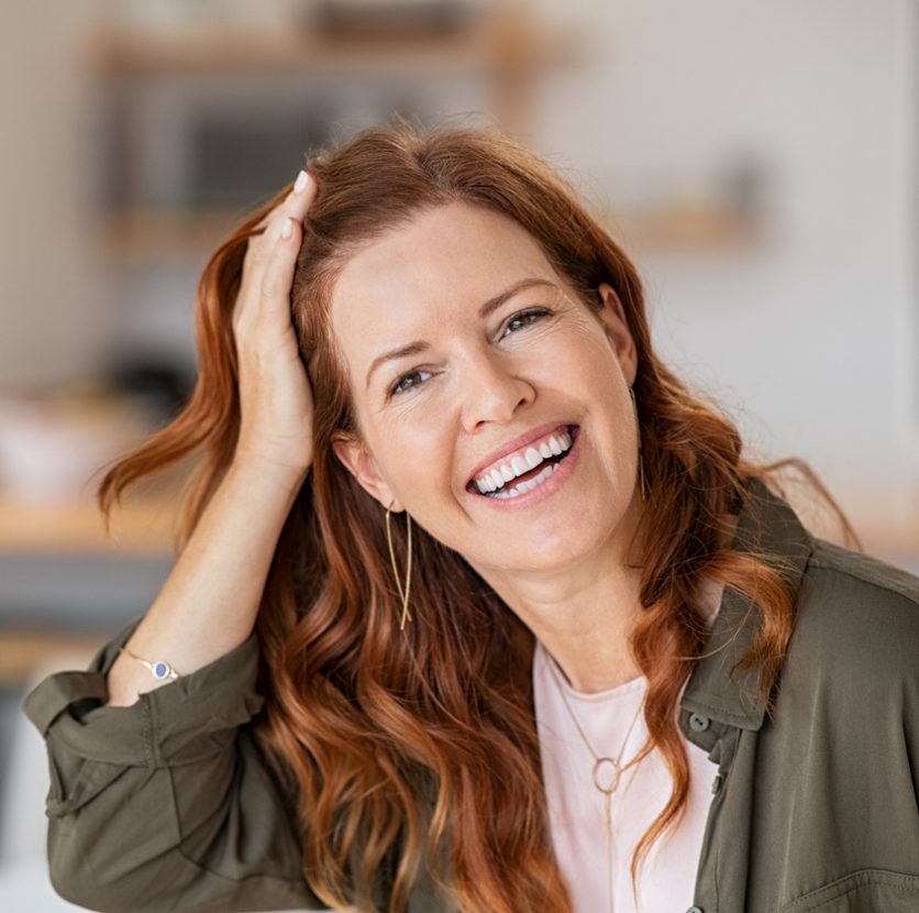 Portrait of cheerful beautiful woman touching hair after anti hair loss treatment. Successful mature woman at home smiling looking at camera. Middle aged redhead woman in casual laughing.