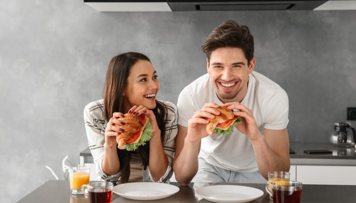 Young and beautiful couple smiling and eating sandwiches on breakfast in kitchen