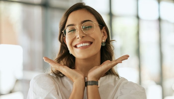 Cheerful business woman with glasses posing with her hands under her face showing her smile in an office. Playful hispanic female entrepreneur looking happy and excited at workplace