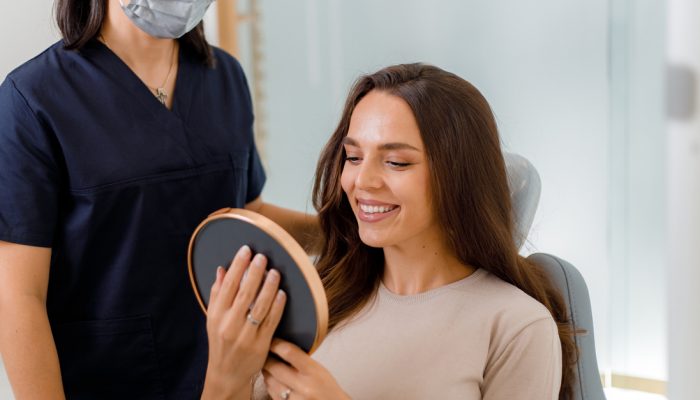 A woman sits in a chair while a woman in a medical uniform holds up a round mirror for her to see. The woman in the chair is smiling and appears satisfied with her reflection.