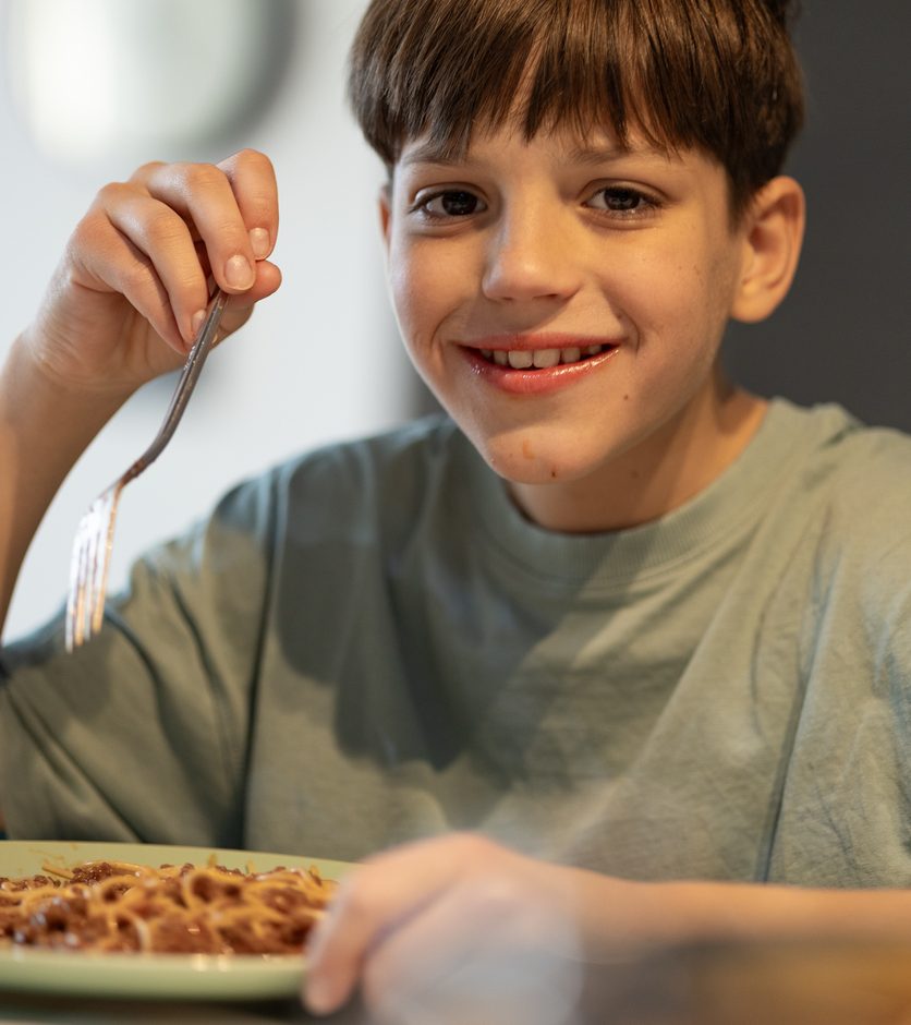 A smiling boy in a green shirt eating spaghetti with a fork