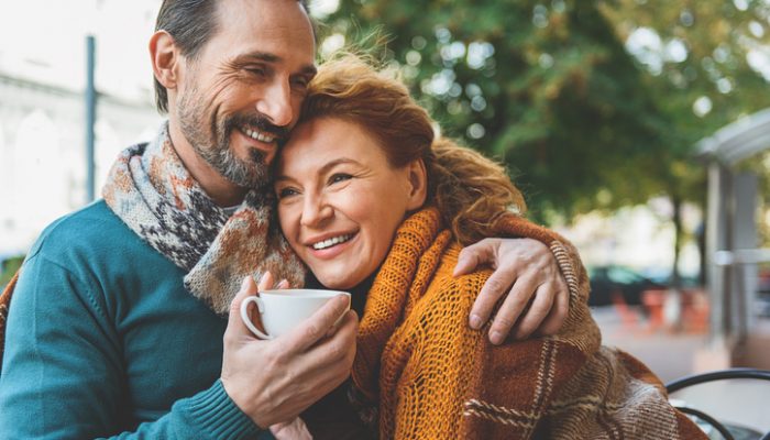 Happy loving couple is dating in cafeteria outdoors. They are sitting covered by blanket and laughing. Man is embracing woman and holding cup